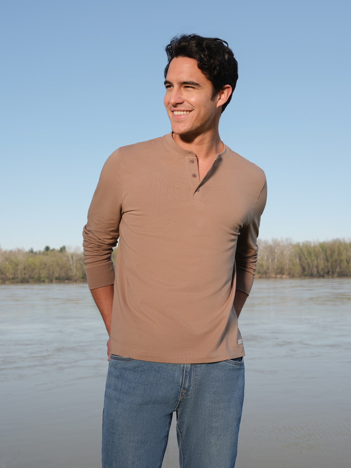 Man wearing a tan long-sleeve henley shirt with button placket and blue jeans standing outdoors.