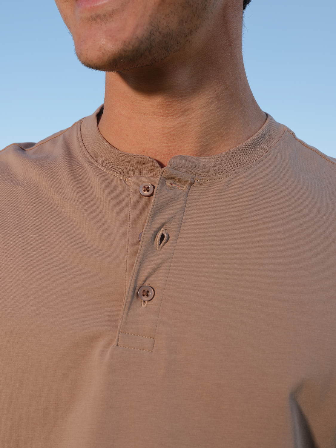 Man wearing a beige Henley shirt with button placket against a clear blue sky.