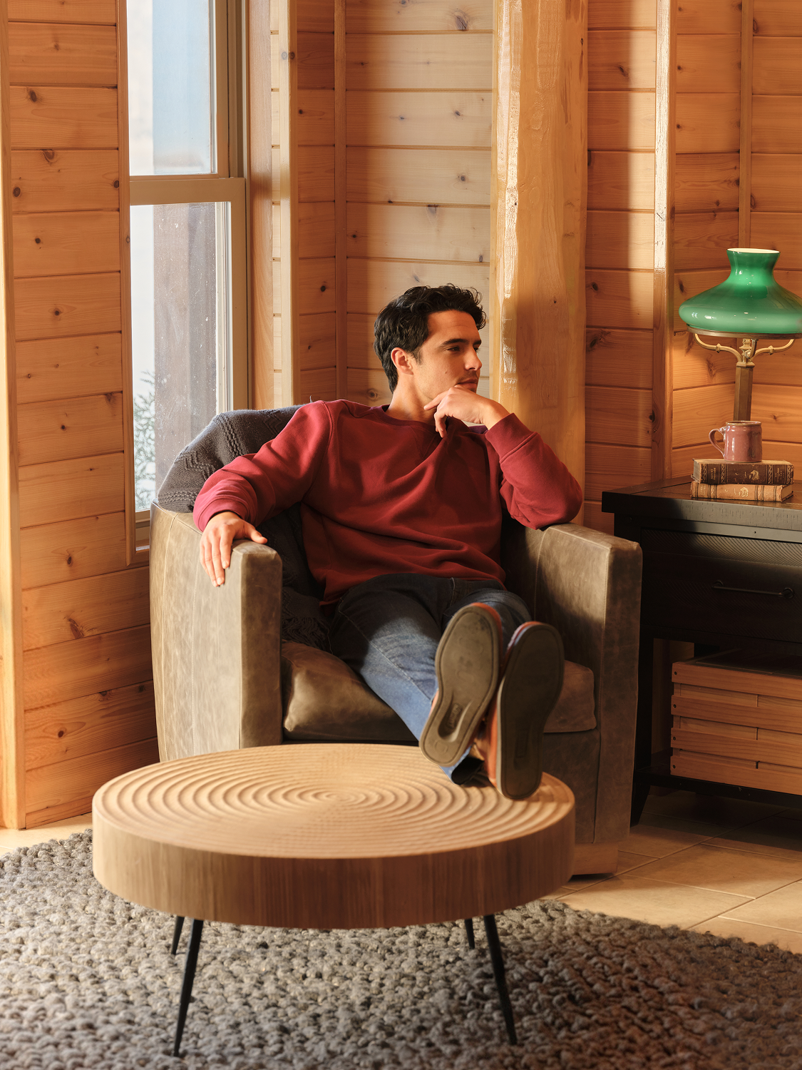 Man wearing a burgundy crewneck sweatshirt and jeans sits relaxed in a beige chair.