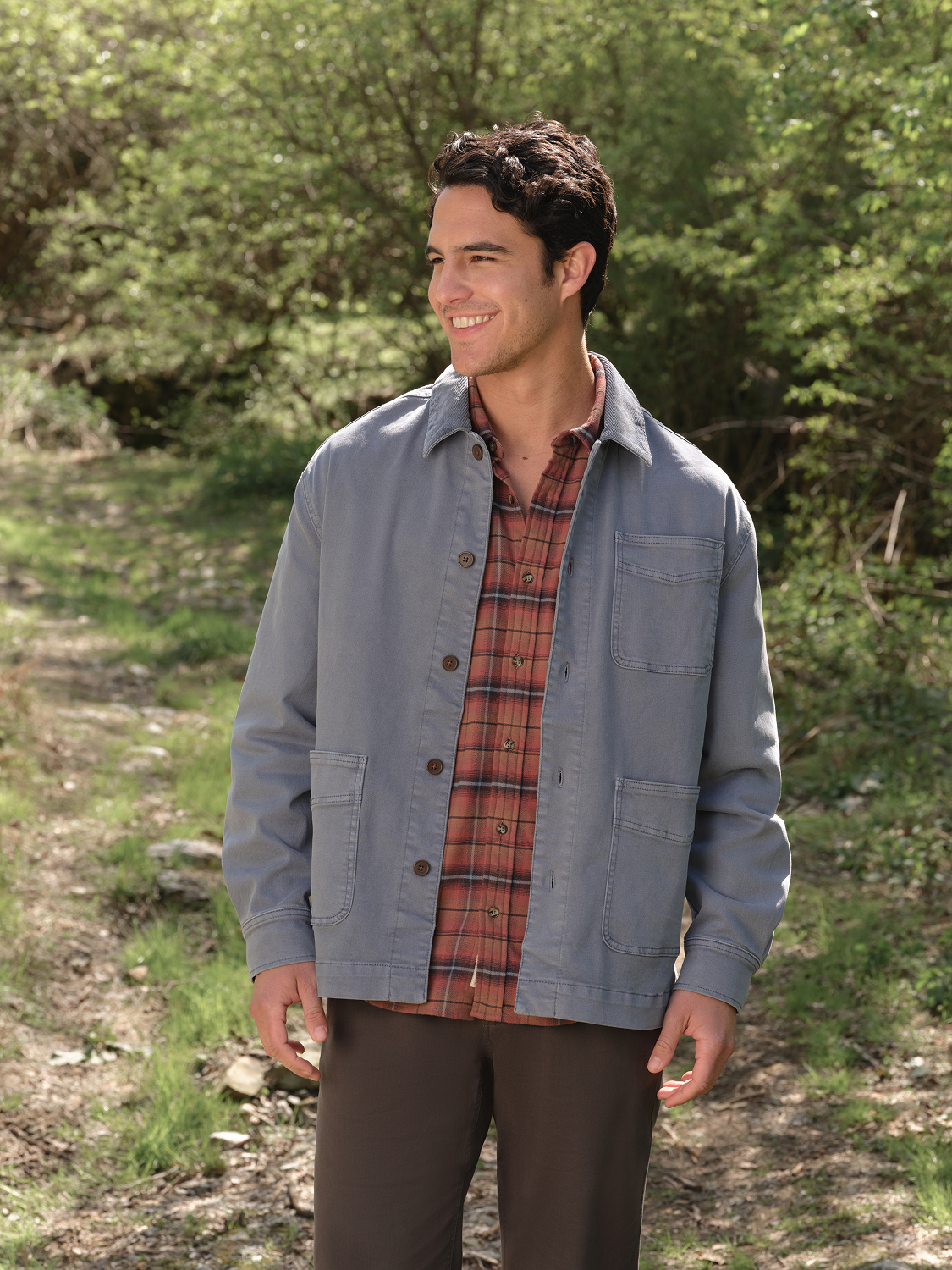 Man wearing a burgundy plaid flannel shirt layered under a gray button-up overshirt outdoors.