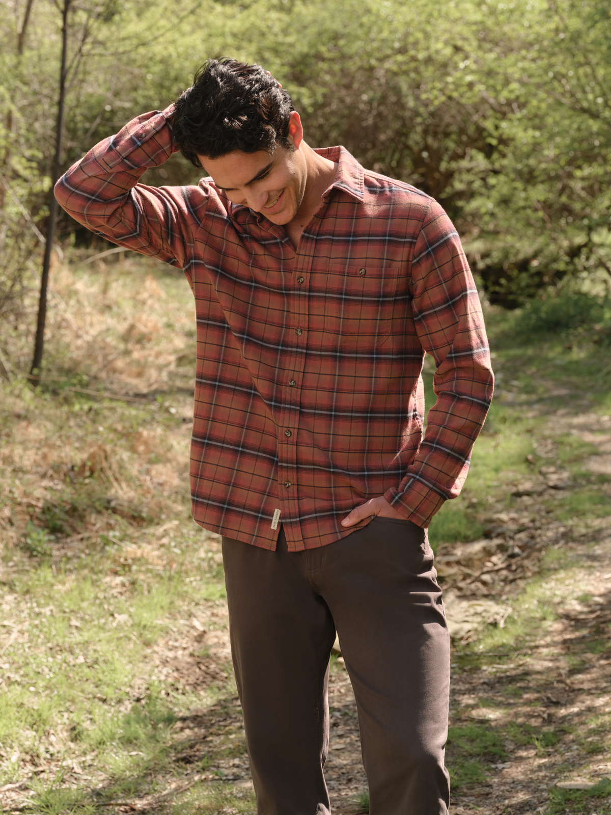 Man wearing a burgundy plaid flannel shirt with dark pants standing on a wooded trail.