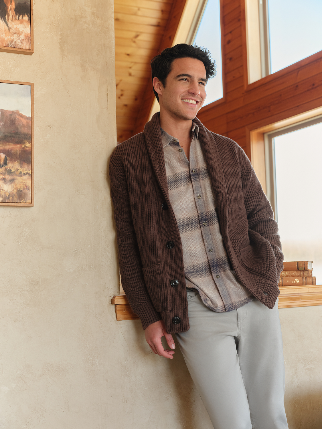 Man wearing a dark brown ribbed cardigan over a plaid shirt and light pants indoors.