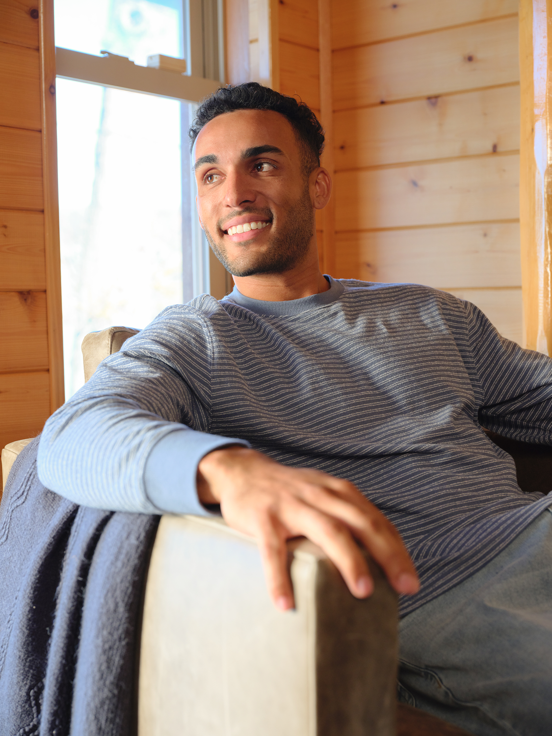 Man wearing a navy striped crewneck sweatshirt sits relaxed indoors near a window.
