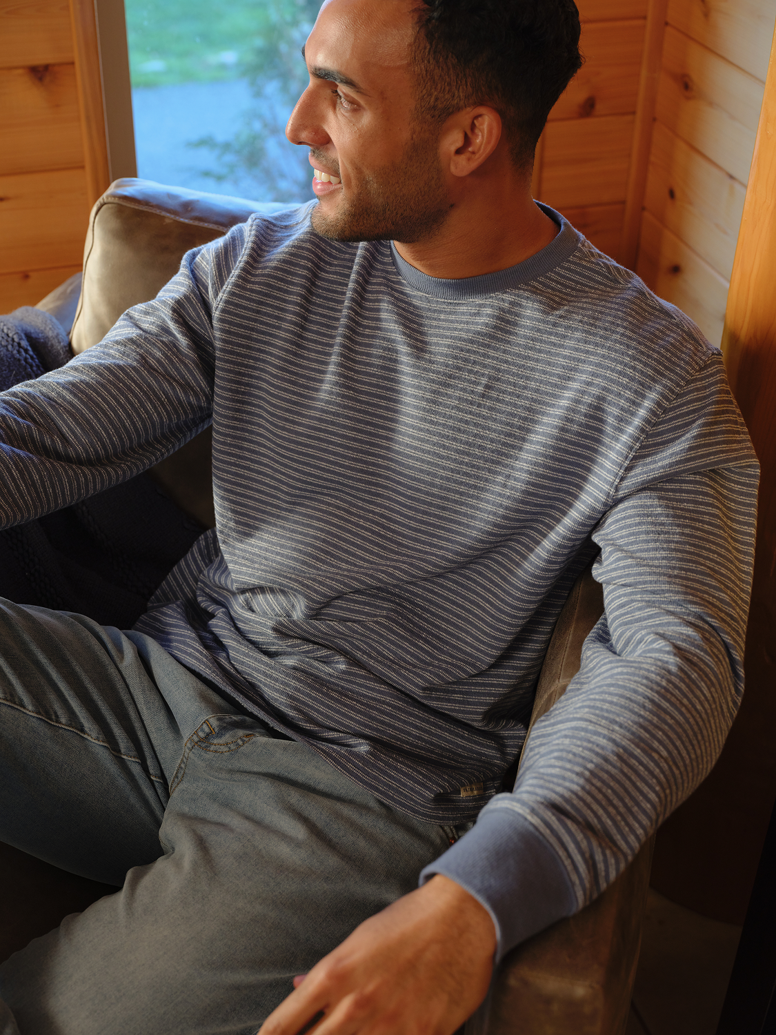 Man wearing a navy striped terry crewneck sweatshirt while sitting on a couch indoors.