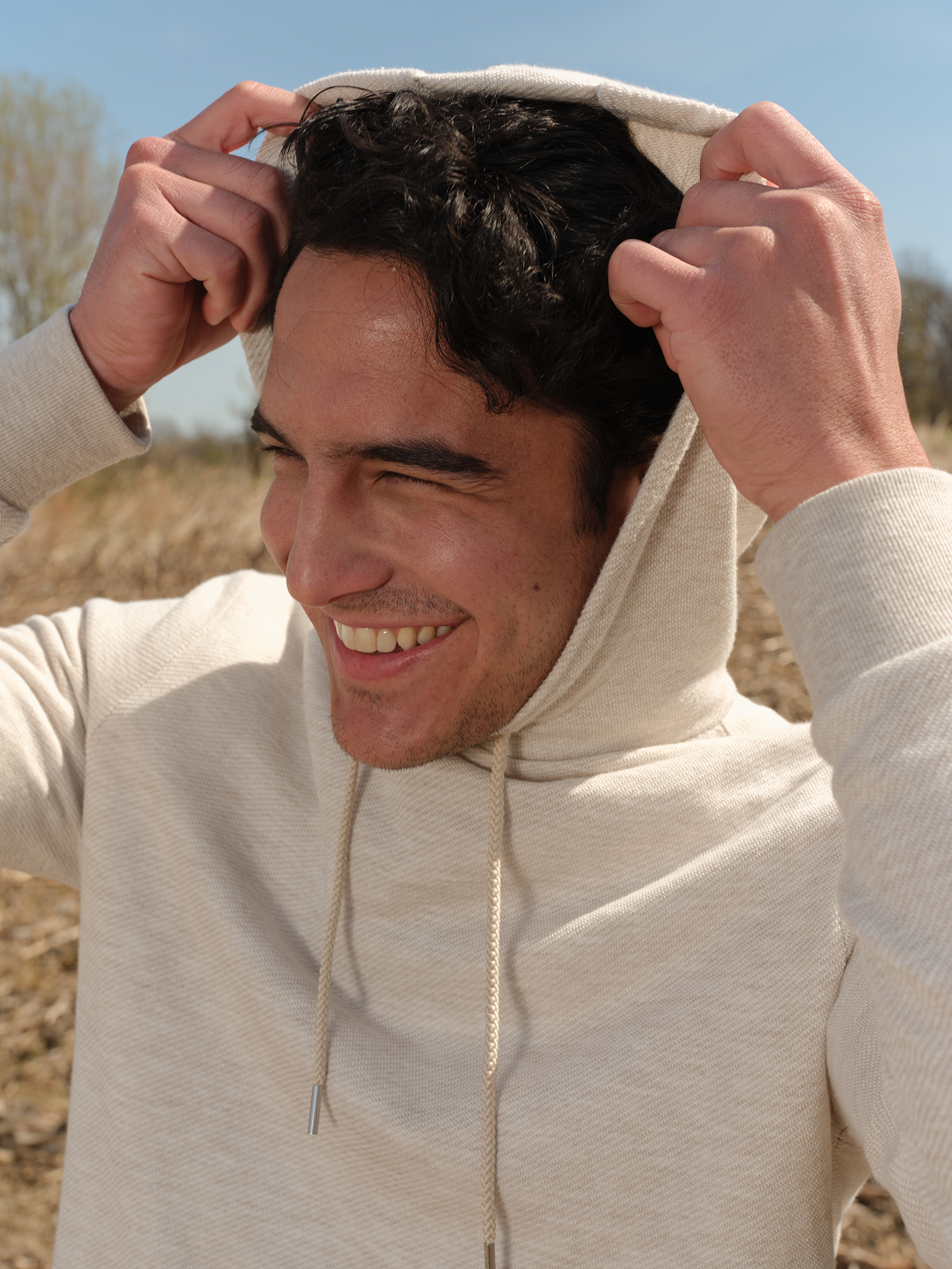 Man wearing a cream-colored hoodie with drawstrings, pulling the hood over his head outdoors.